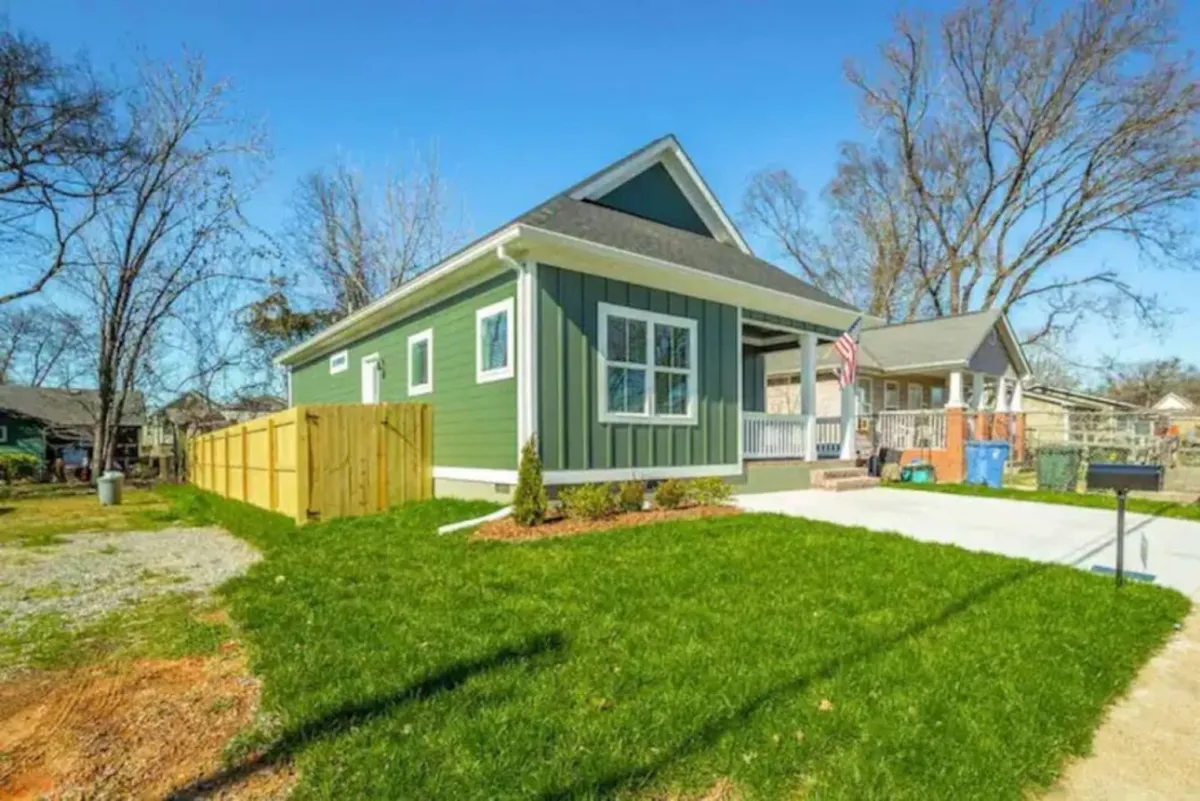 An exterior view of a well-maintained home with a green façade, white trim, a wide front porch, an American flag, and a manicured lawn. The scene is lively and welcoming, suggesting a base for families and groups setting out to explore the city.