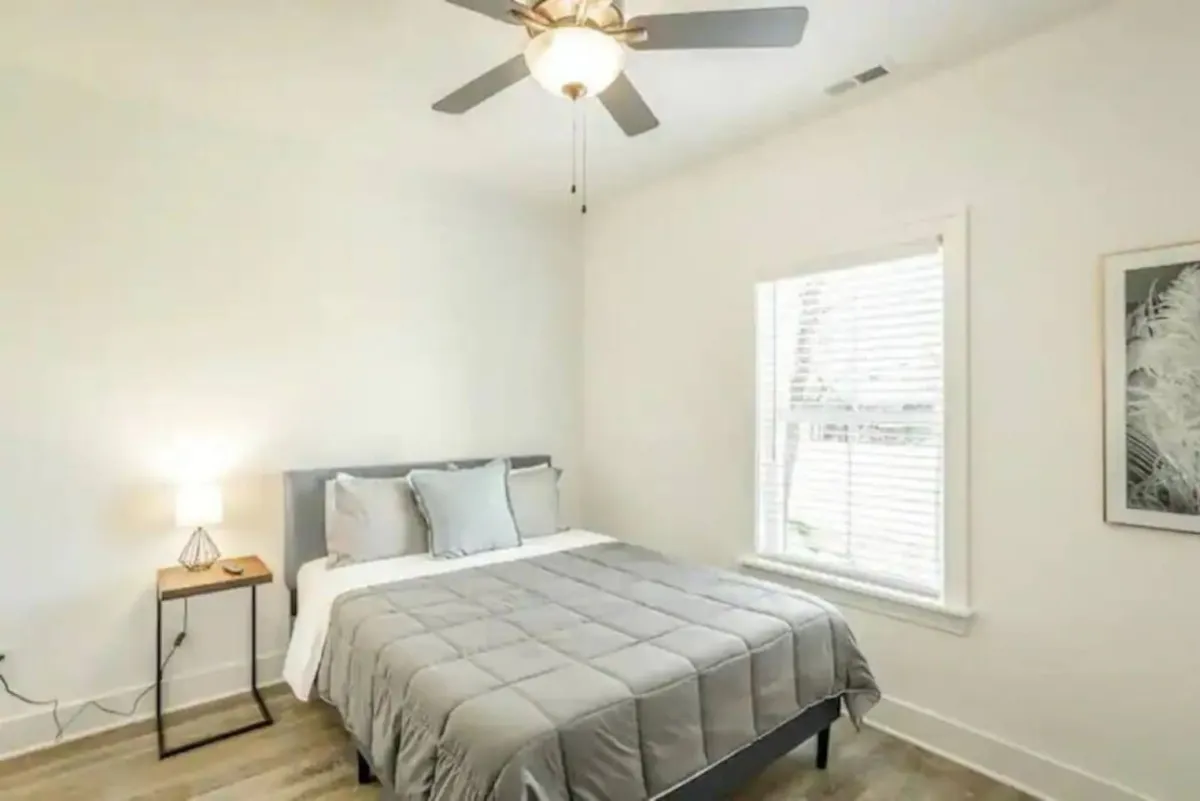 This image features a tranquil bedroom with a plush bed, a cozy reading nook by the window, a nightstand with a lamp, and a ceiling fan overhead. The soft lighting and serene setup invite relaxation and restful sleep.