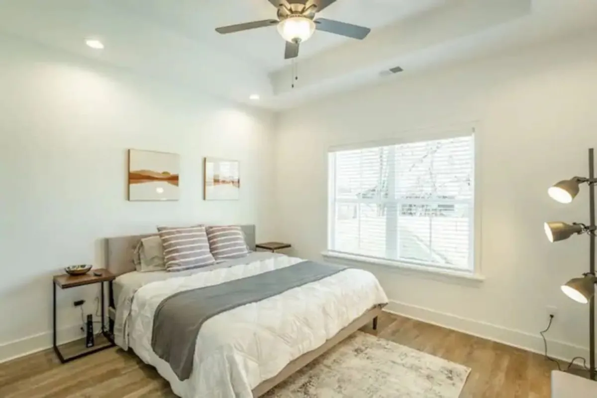 A tranquil bedroom scene with a neatly made bed, window, lamp, and artwork, evoking a sense of relaxation and comfort. The soft lighting and thoughtful furnishings suggest a restful atmosphere, perfect for recharging after late-night adventures.
