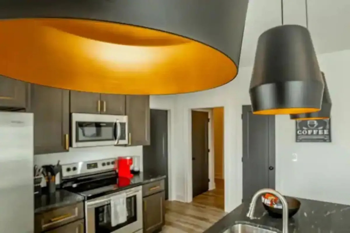 A stylish kitchen featuring large pendant lights with orange interiors, stainless steel appliances, dark cabinetry, and white countertops. The scene hints at cooking and entertaining, with ample space around the counters and a cozy, welcoming mood.