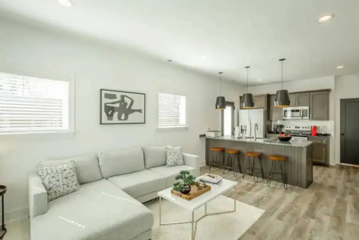 A modern, spacious living area featuring a comfortable sofa, a kitchen island in the background, striking light fixtures, and contemporary artwork on the wall. The open layout invites both relaxation and social gatherings, with sightlines connecting the lounge to the kitchen.