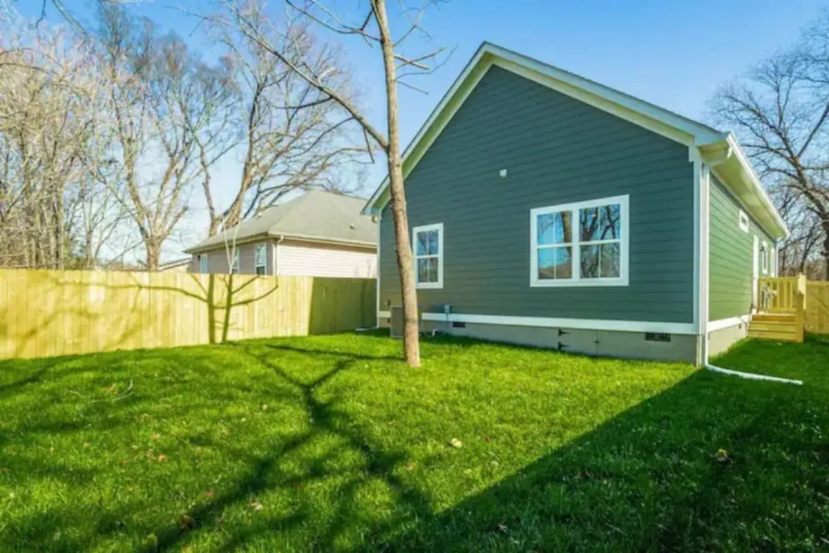 A cheerful exterior shot showing the green siding of the house, a well-maintained lawn, and a tree casting soft shadows. The welcoming mood suggests sunny afternoons, barbecues, and enjoying nature right outside your door.