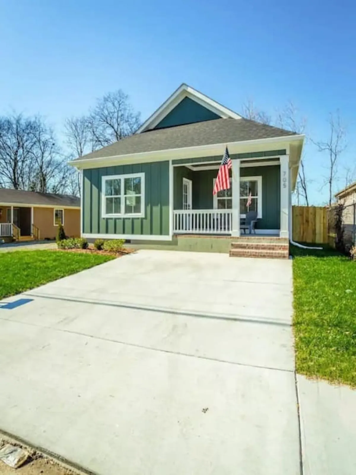 A sunny exterior shot emphasizing the front of the home, green siding, white trim, and American flag. The porch looks ready for relaxing with morning coffee or greeting neighbors after a day out.