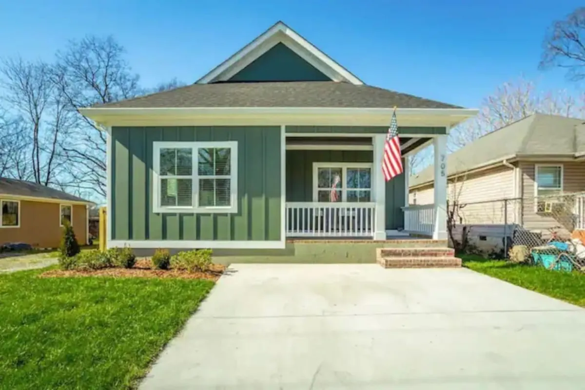 This image shows a classic front porch with green siding, an American flag, and a welcoming, well-kept appearance. The porch has seating and the yard is neatly maintained, suggesting a home that sits pleasantly in its neighborhood and is ready for outdoor relaxation or greeting neighbors.