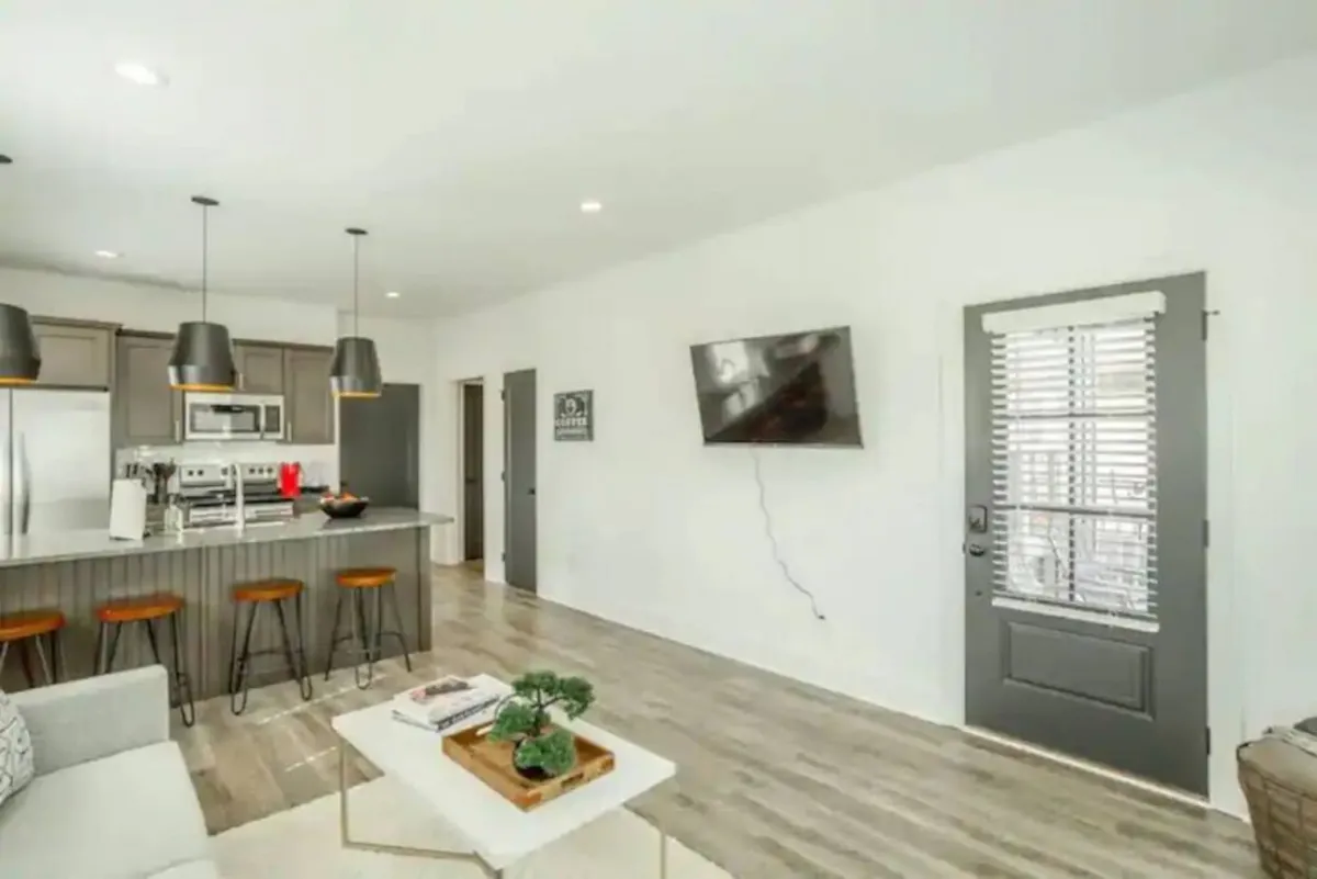 A bright, inviting view of the living area with a kitchen island, comfortable seating, a television visible from the lounging space, and a glimpse of the entryway. The arrangement is both practical and welcoming, highlighting multiple entertainment options.