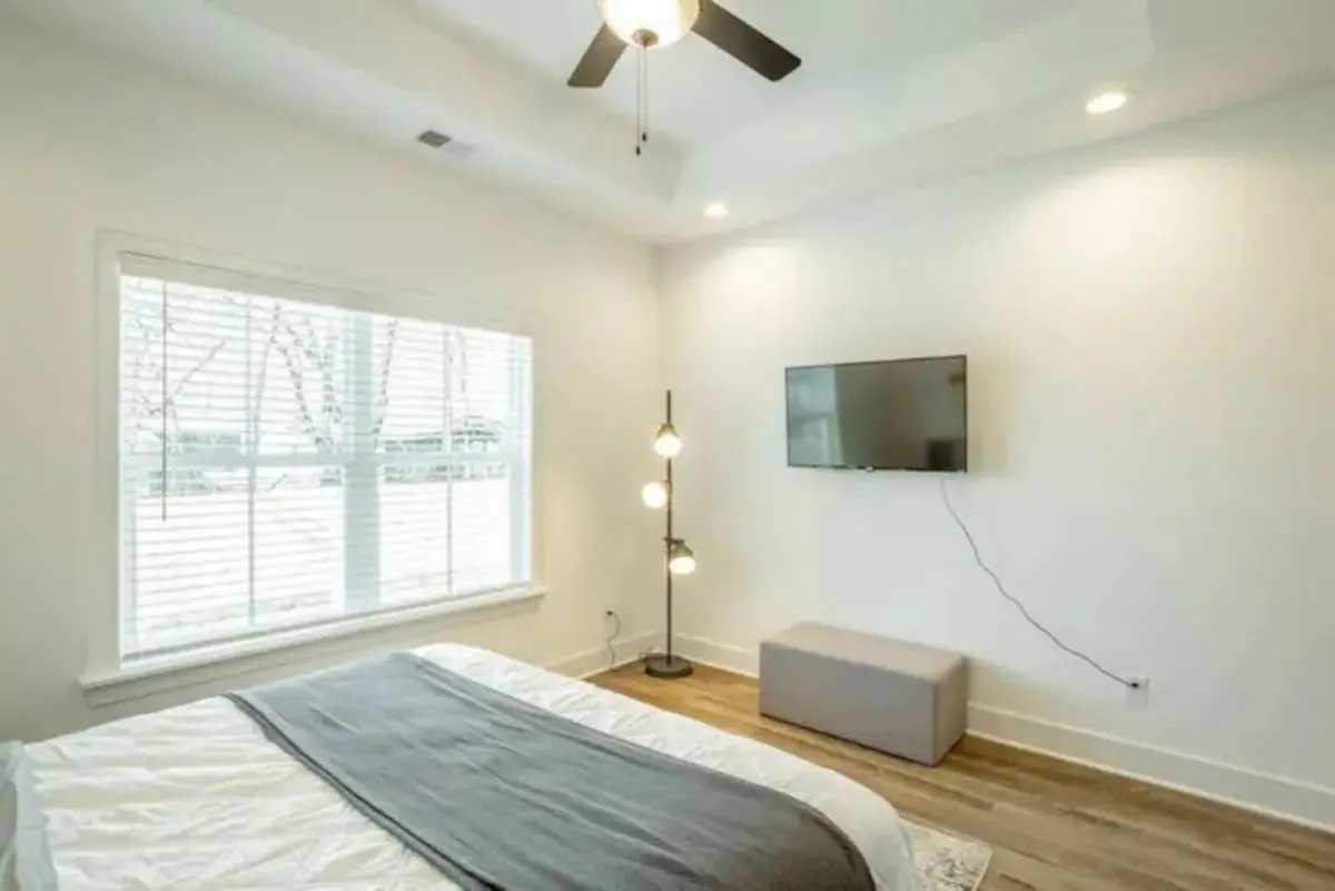 A bright, inviting bedroom with a plush bed topped with a gray throw, a lamp on the nightstand, a large window letting in natural light, and a wall-mounted smart TV. The space feels modern and comfortable, ideal for relaxing or enjoying your favorite show from bed.