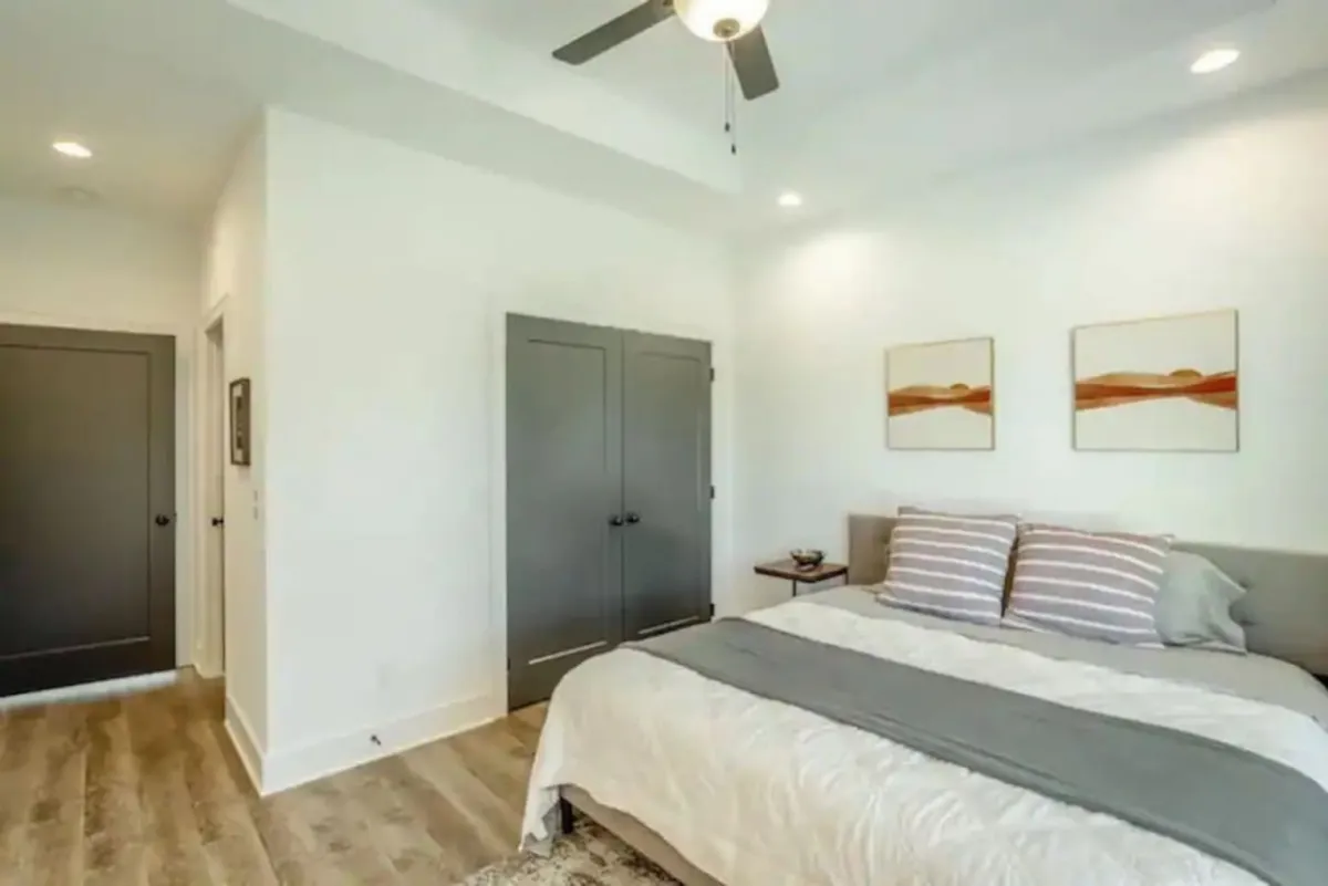 A tranquil bedroom scene featuring a bed with a soft gray throw, two pieces of framed artwork above the bed, and dark closet doors. Gentle daylight streams in, creating a peaceful mood ideal for reading, napping, or simply unwinding in private.