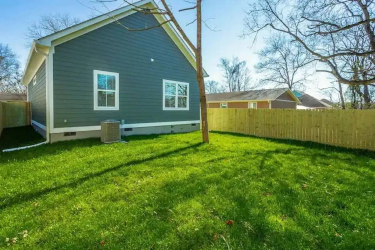 This image provides a clear view of the securely fenced yard bordered by green grass and the gray siding of the house. The fence is prominent, with open lawn spaces ideal for children and pets to play safely. The overall atmosphere is peaceful and inviting, suggesting a private outdoor haven.